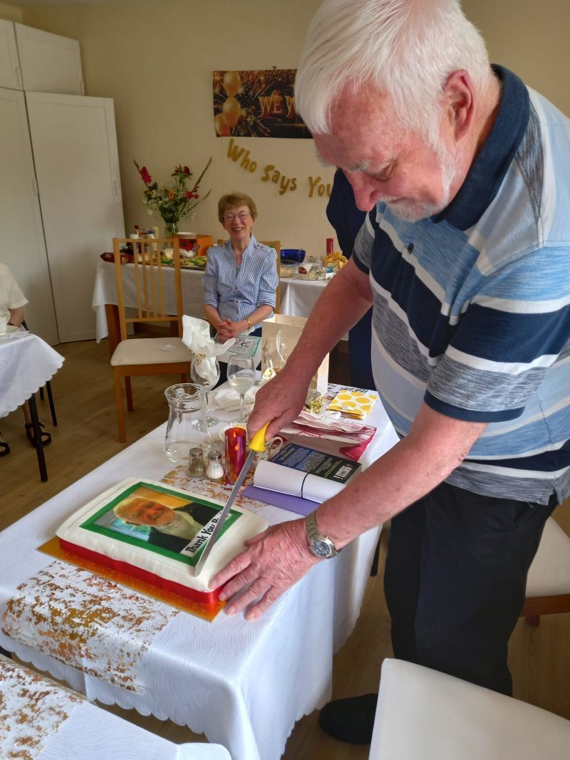 Man cutting a cake with a picture on top, at a party; woman watching at a table.