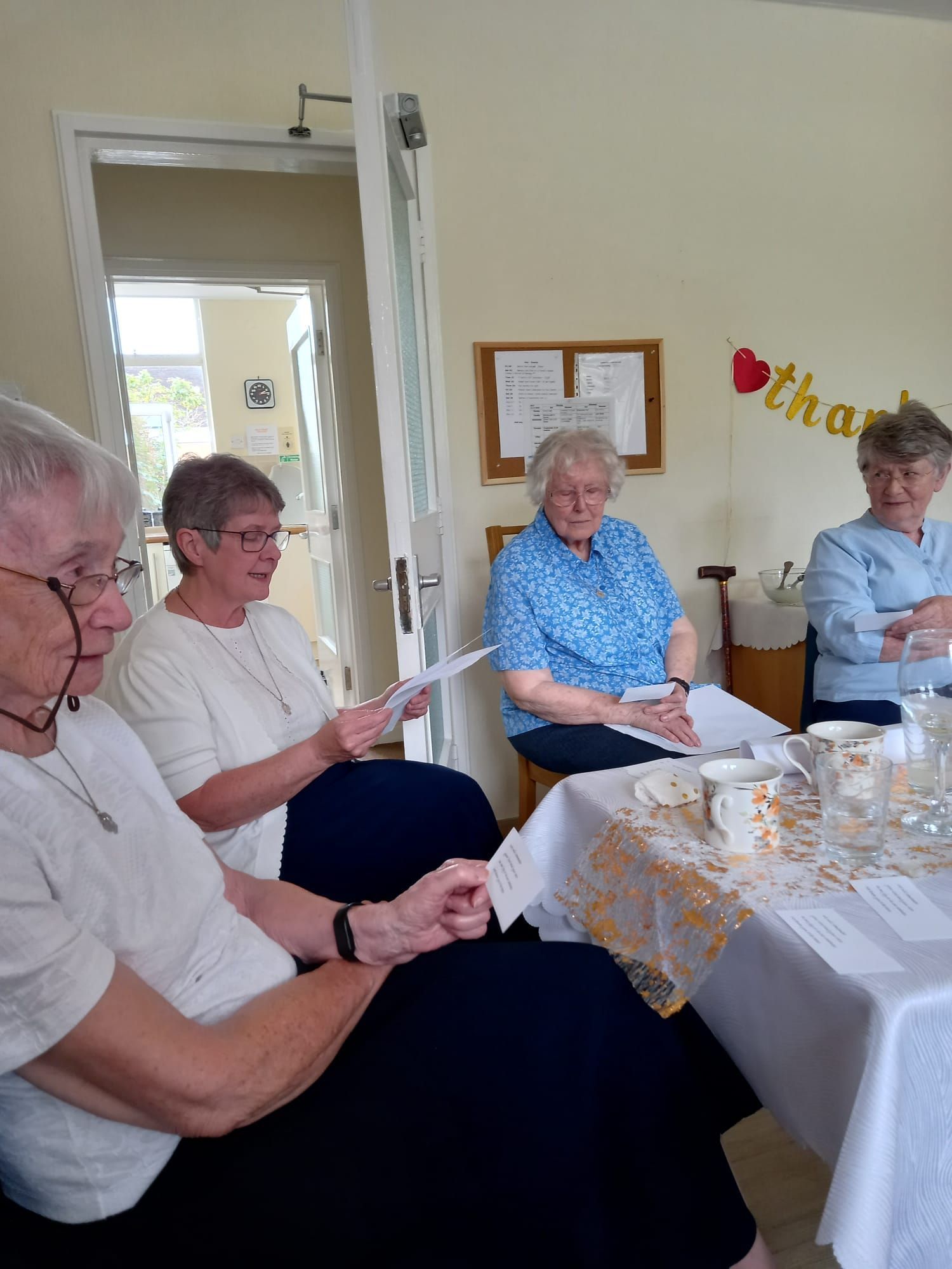 Four elderly women sitting at a table, possibly in a care home, reading papers and talking.