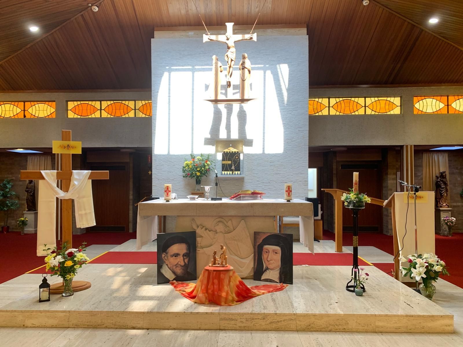 Church interior with altar, crucifix, portraits, and flowers. Sunlight streams through the window.
