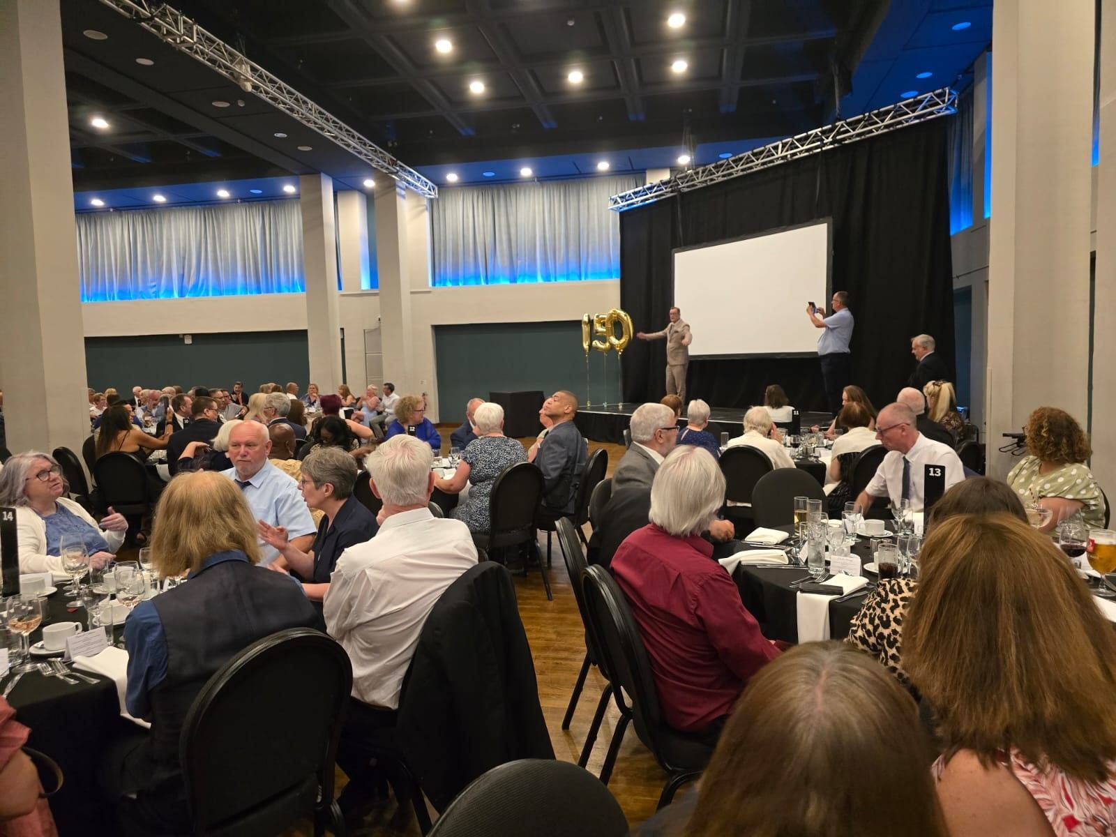 Guests at a banquet, focused on a stage with a screen. People in formal attire at tables.