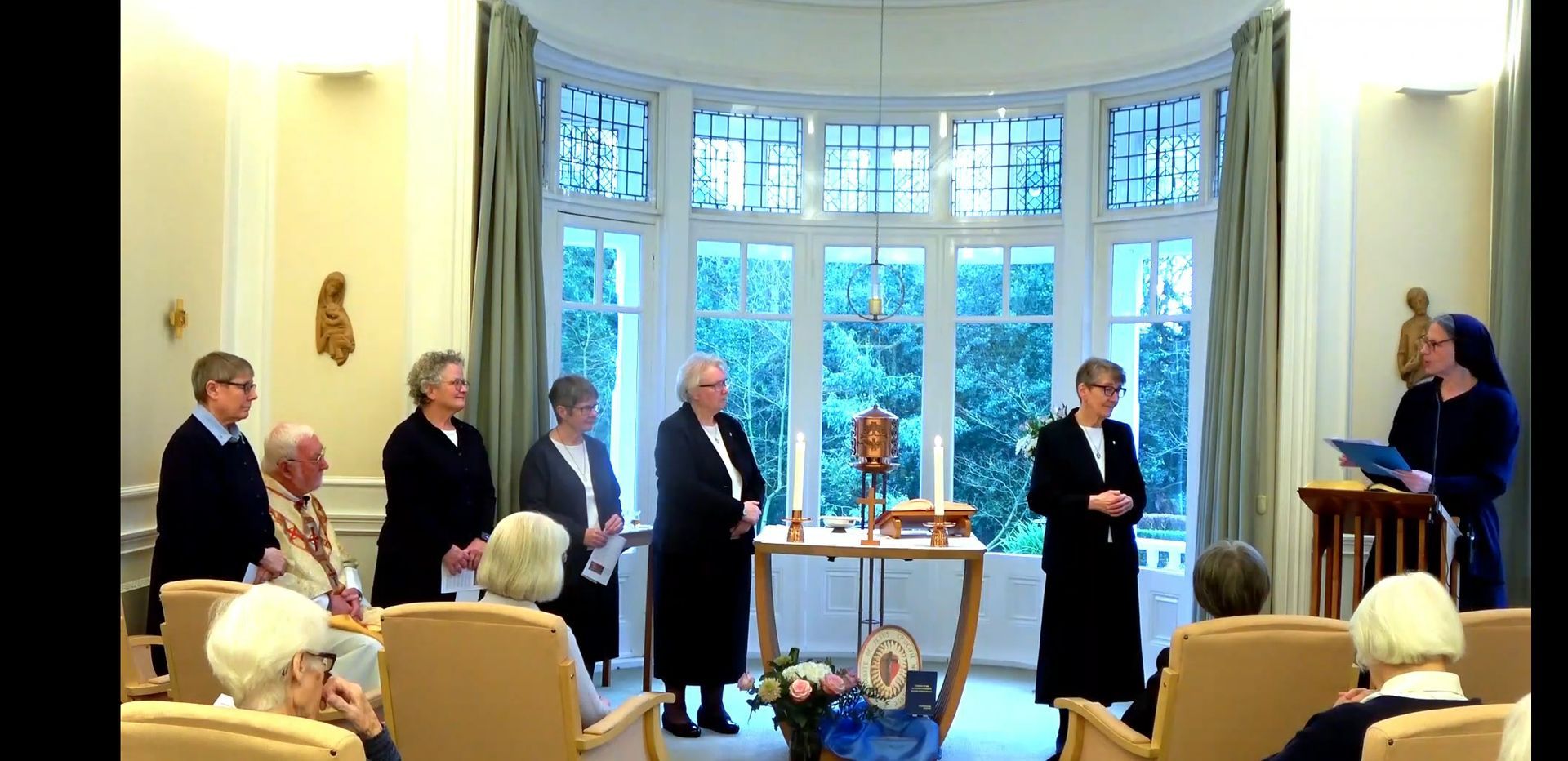 Nuns and elderly people gathered in a room, centered around a small altar, some facing the window.
