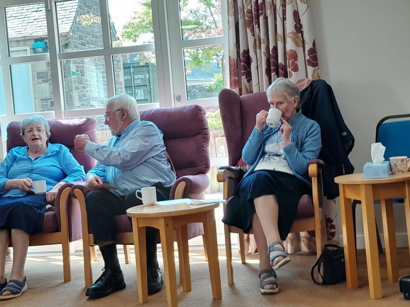 Three older adults in a well-lit room with coffee. Two women and a man are seated, talking and gesturing.