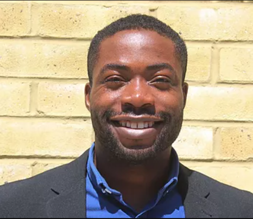 A man in a suit and blue shirt is smiling in front of a brick wall.