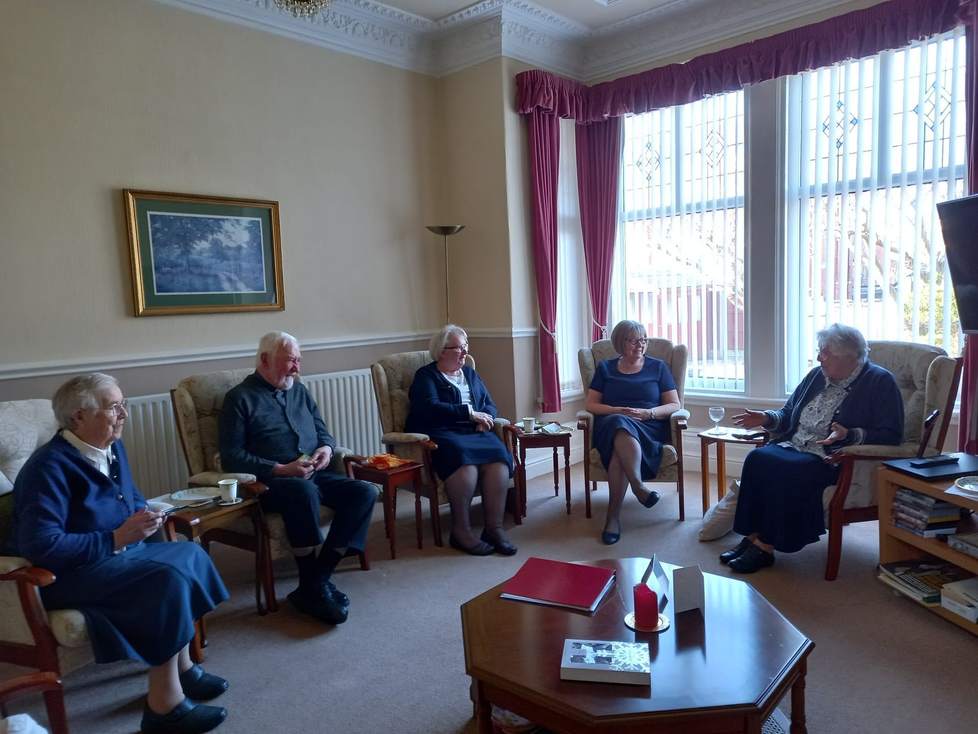 Five seated elderly people conversing in a living room. They are in armchairs around a coffee table. Bright sunlight.