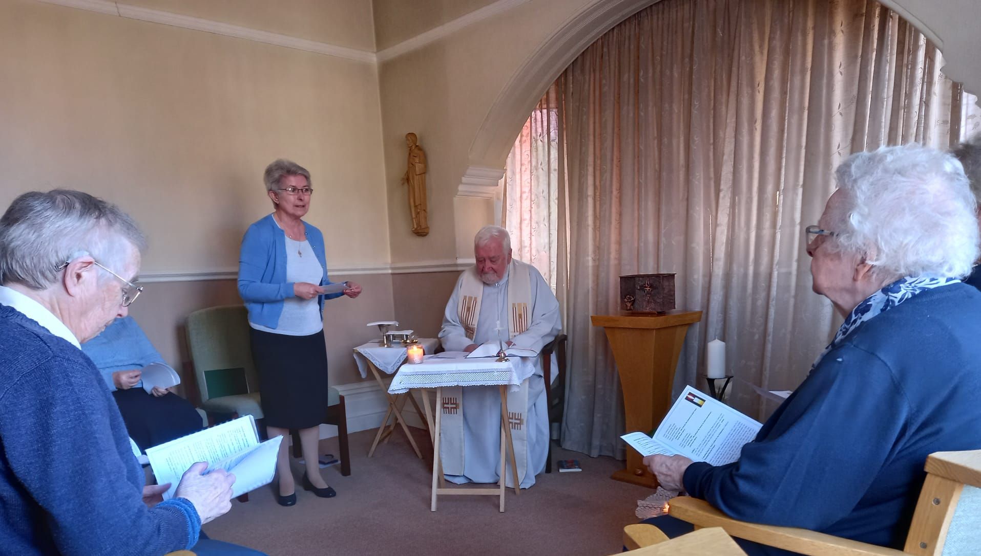 A priest leading a service in a room with several older adults; a woman speaks at the lectern.