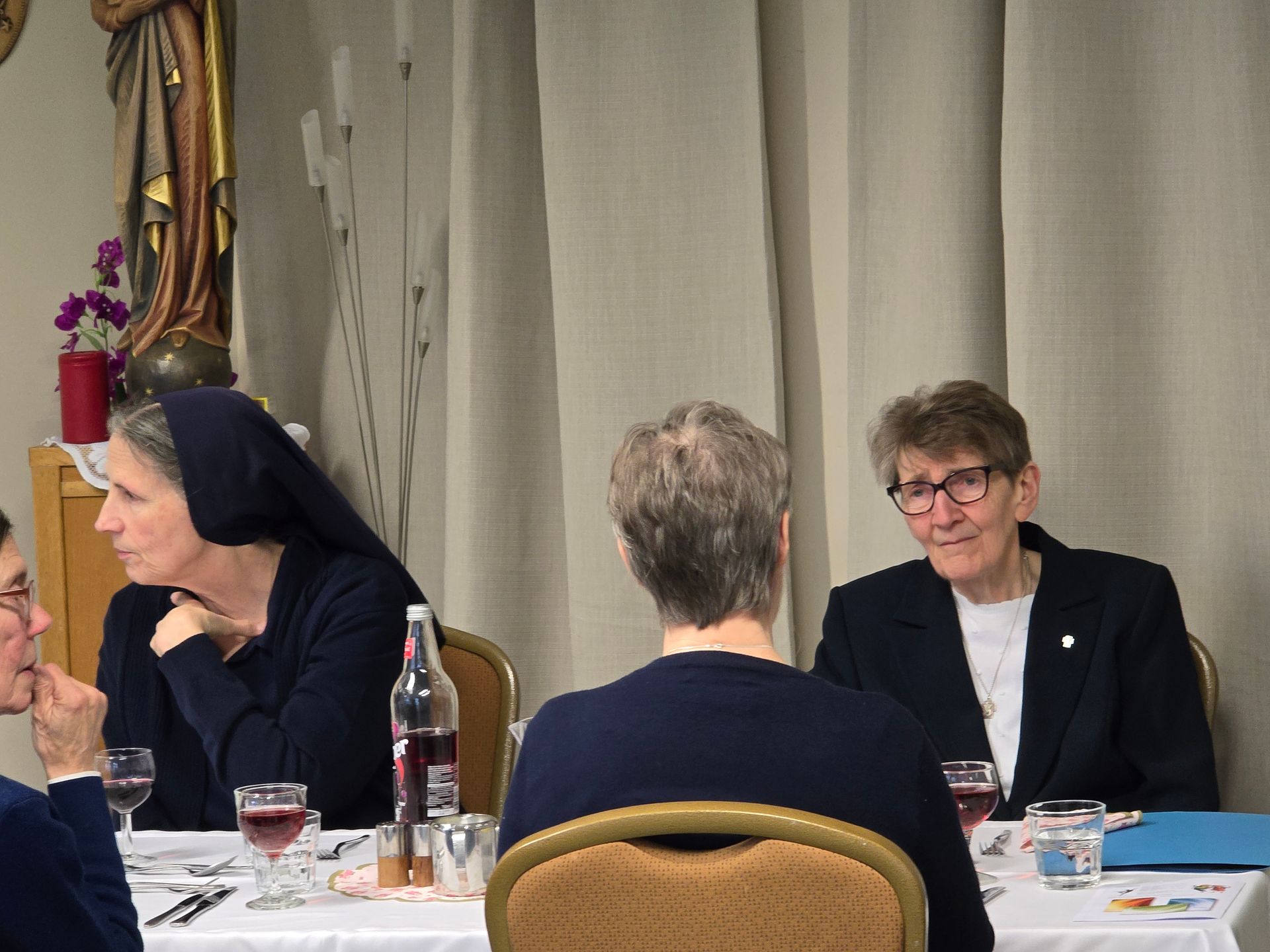 People, including nuns, seated at a table, talking, drinking wine, in a dining area.