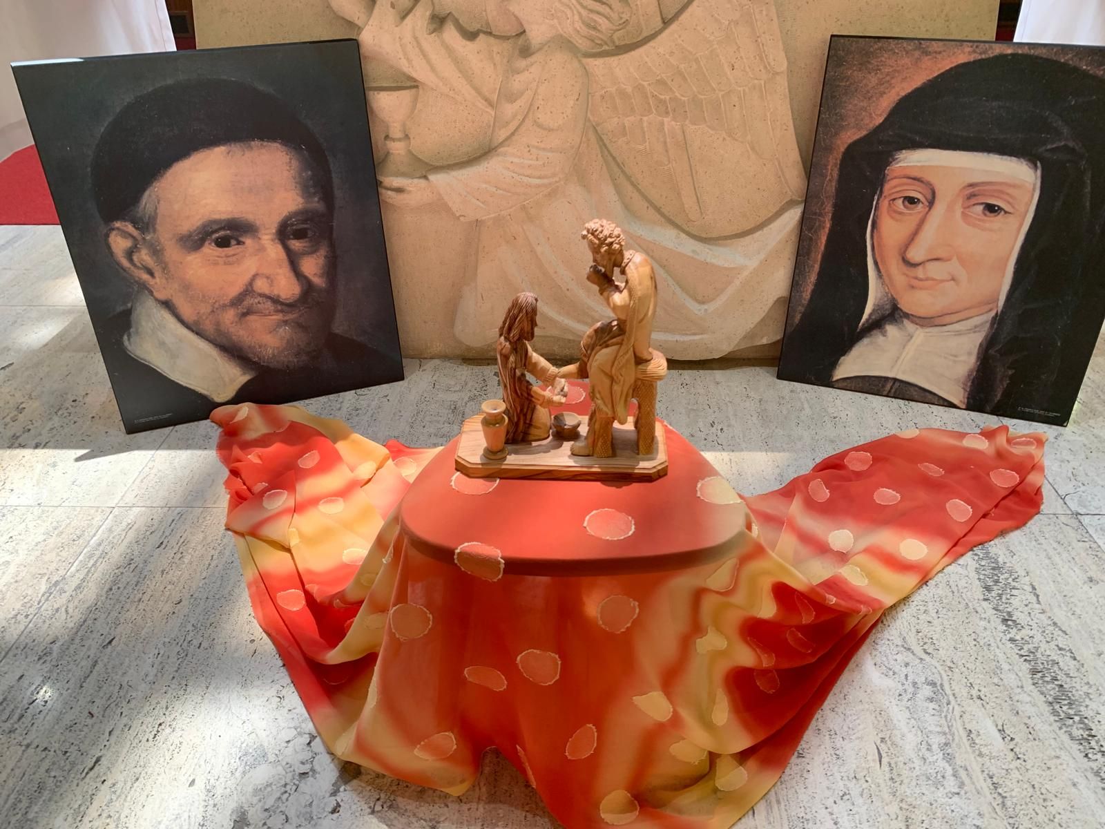 Portraits flank a covered container topped with a statue; all on a colorful cloth.