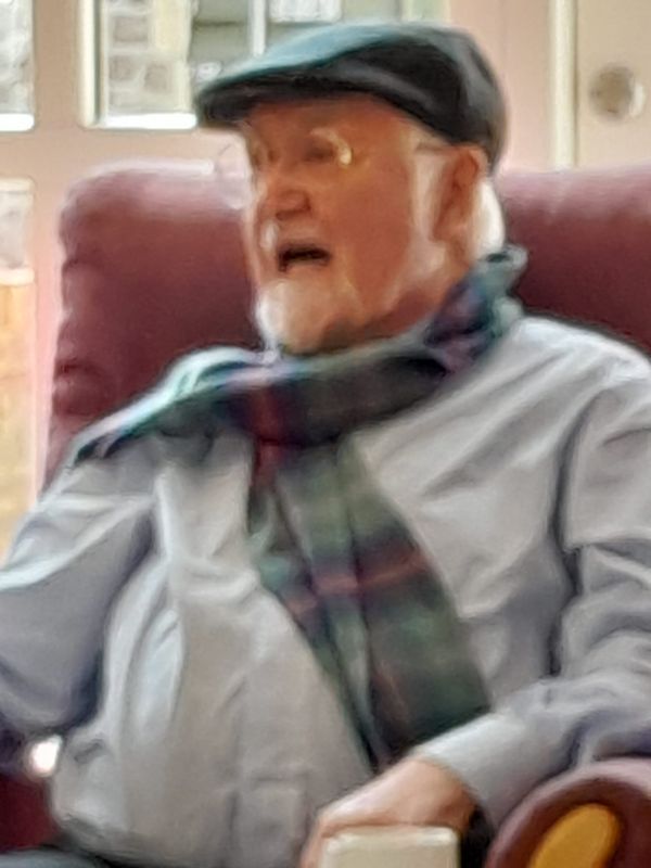 Elderly man wearing a flat cap and scarf, sitting in a chair, talking.