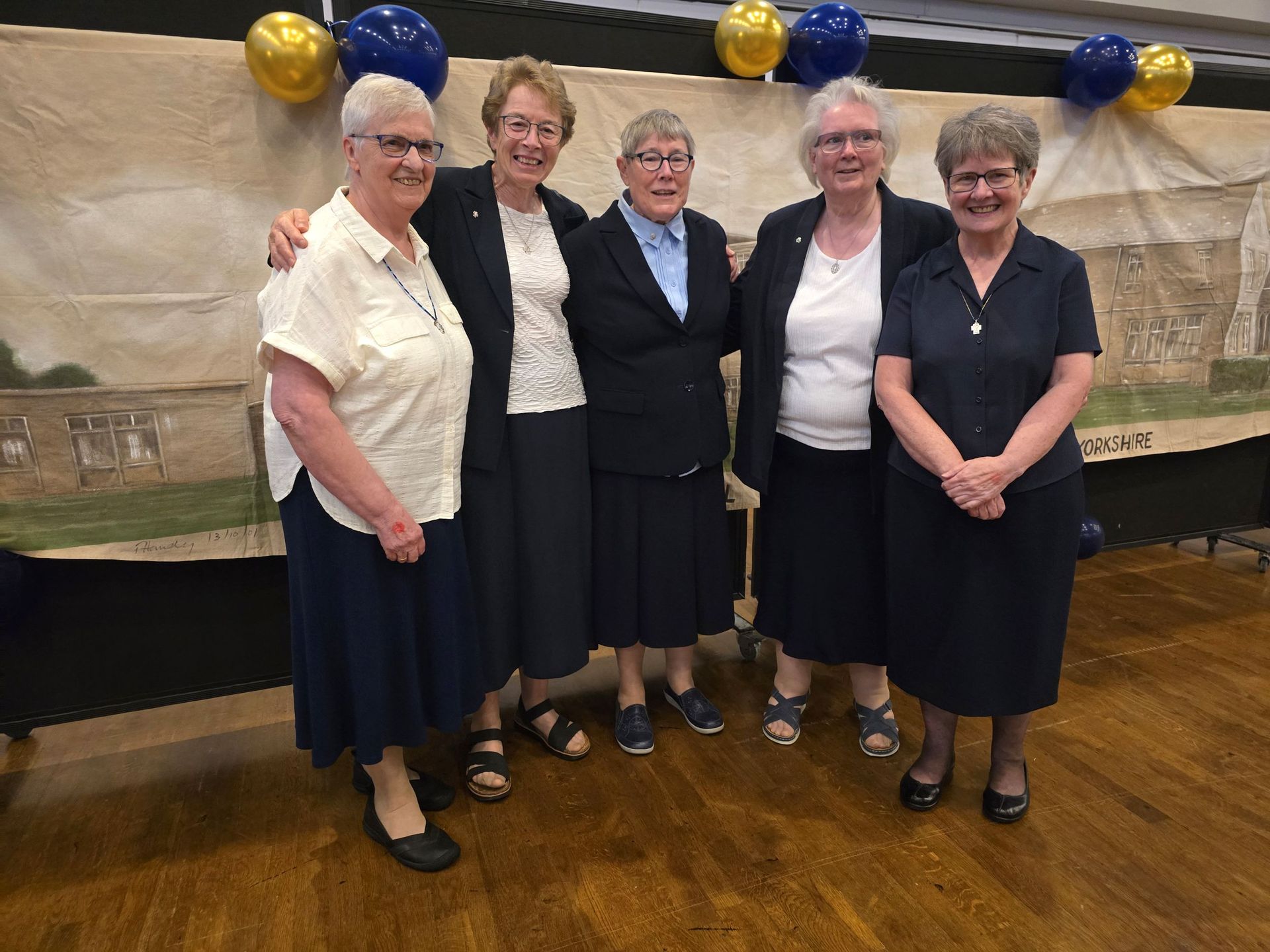 Five women in dark skirts and jackets pose in front of a banner, smiling. Blue and gold balloons.