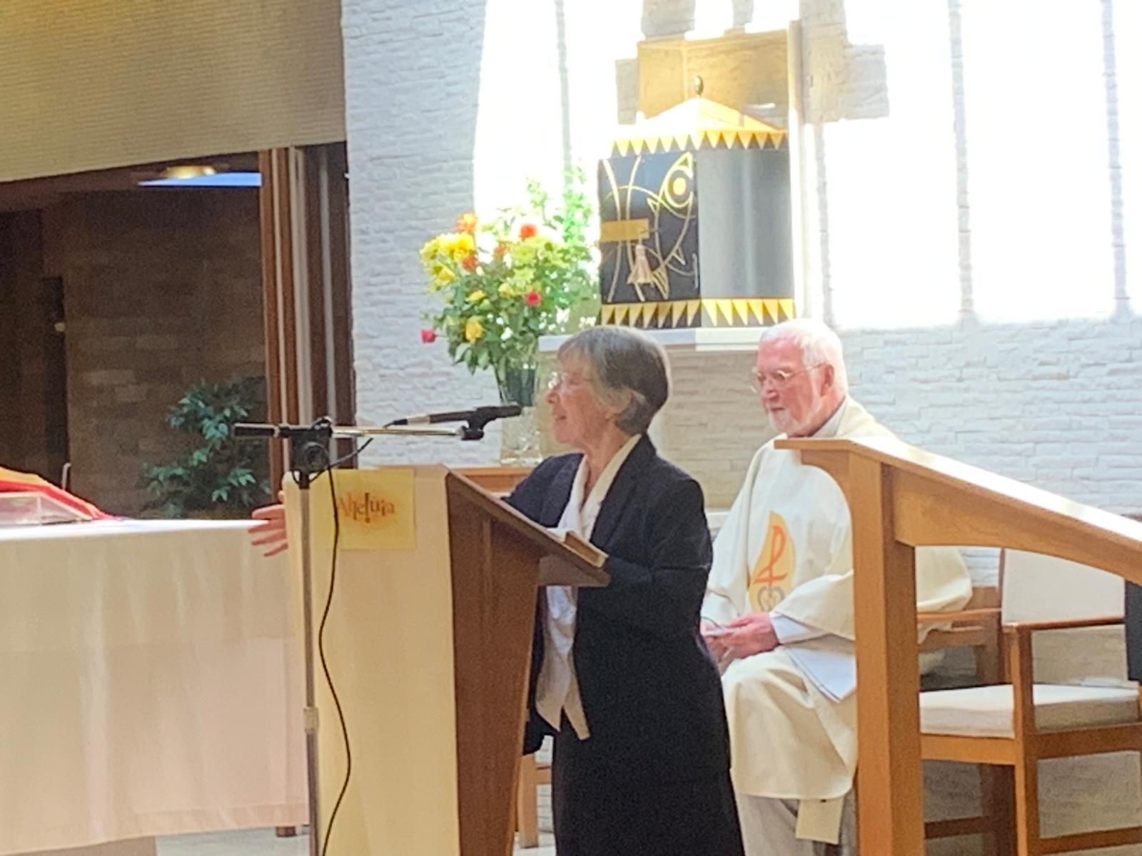 Woman speaking at a podium in a church, with a man in religious garb seated beside her.