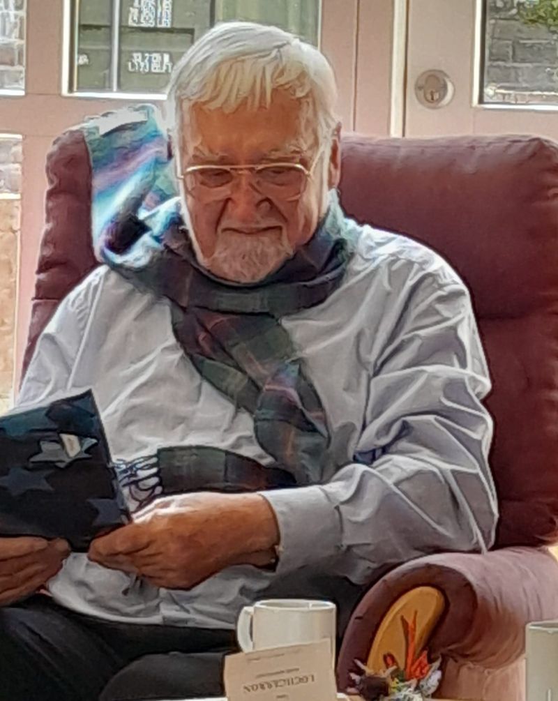 Older man with white hair and glasses seated in a chair, holding a gift and wearing a scarf.