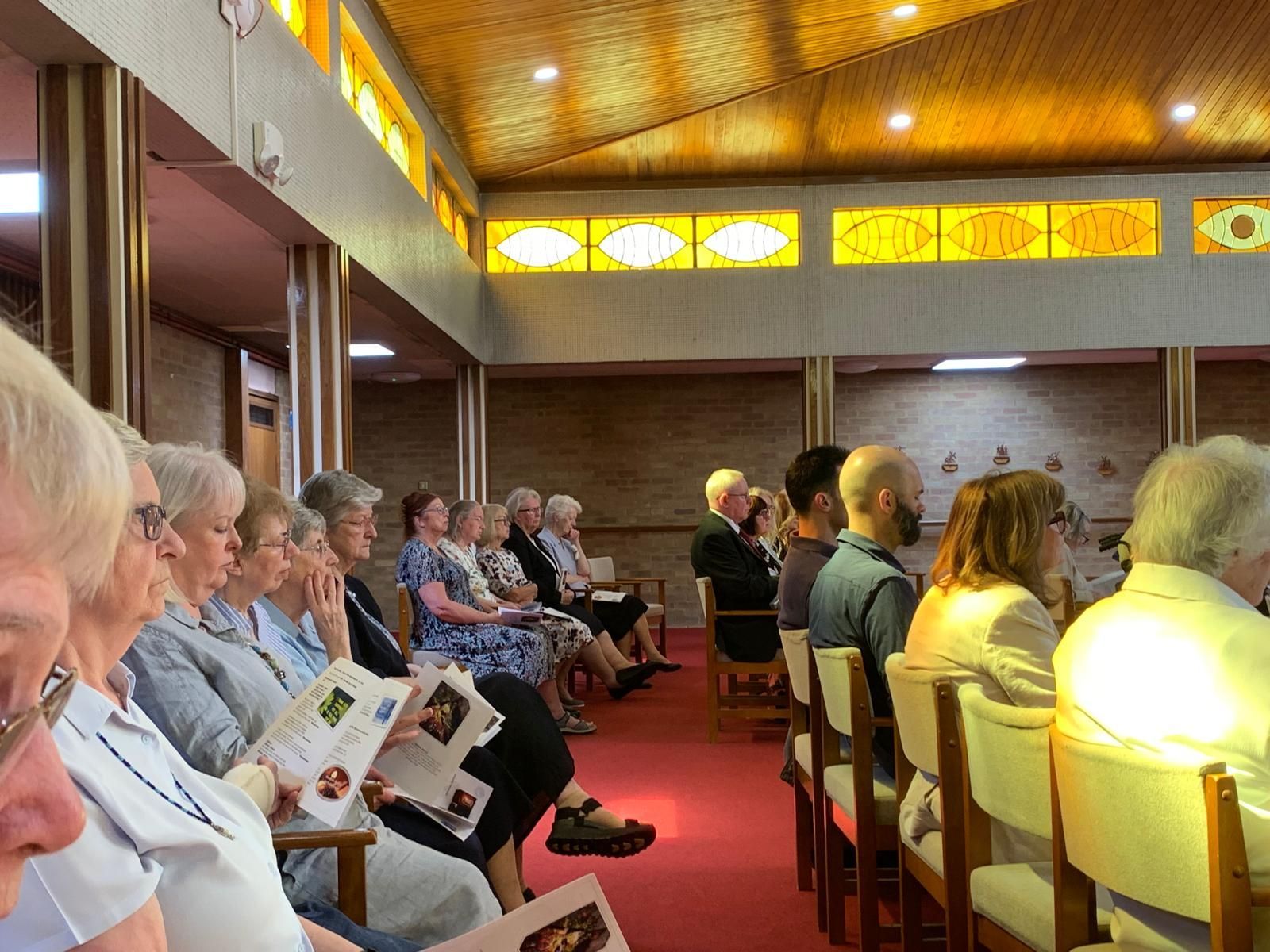 People seated in pews during a service, likely a church. Warm lighting, red carpet, stained-glass windows.
