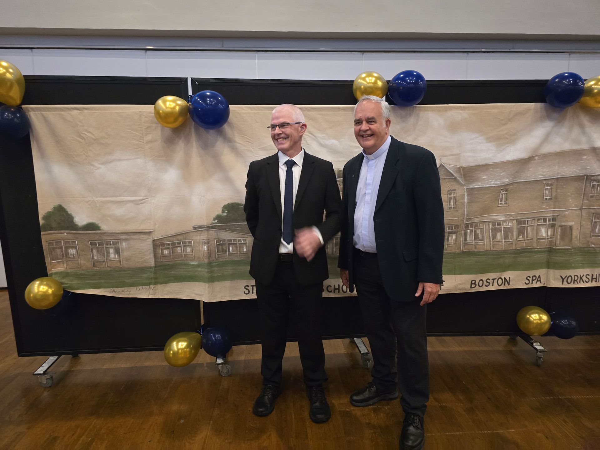 Two men in suits stand in front of a banner with a building illustration and balloons.