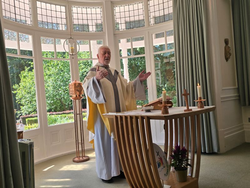 Priest in white vestments giving a sermon, standing at a wooden altar in front of a large window.