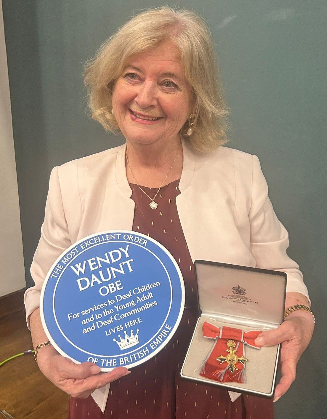 Woman holding blue plaque and medal, smiling, wearing light jacket and burgundy dress.