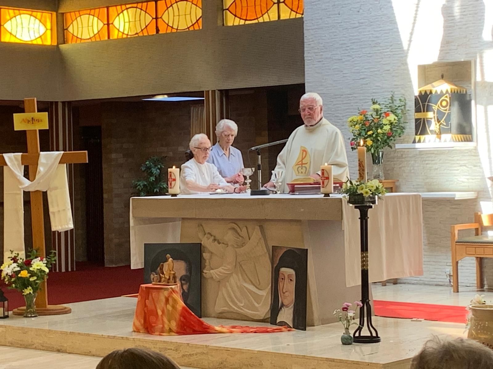 Priest leading a service with two nuns. Portraits, cross, and stained-glass windows in a church.