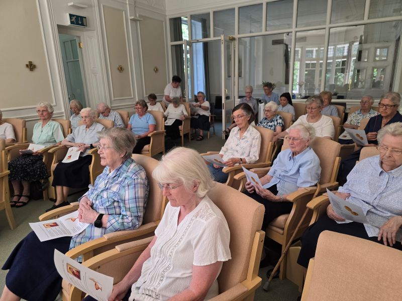 A group of elderly people seated indoors, listening attentively. Some hold papers. Sunlight streams in.