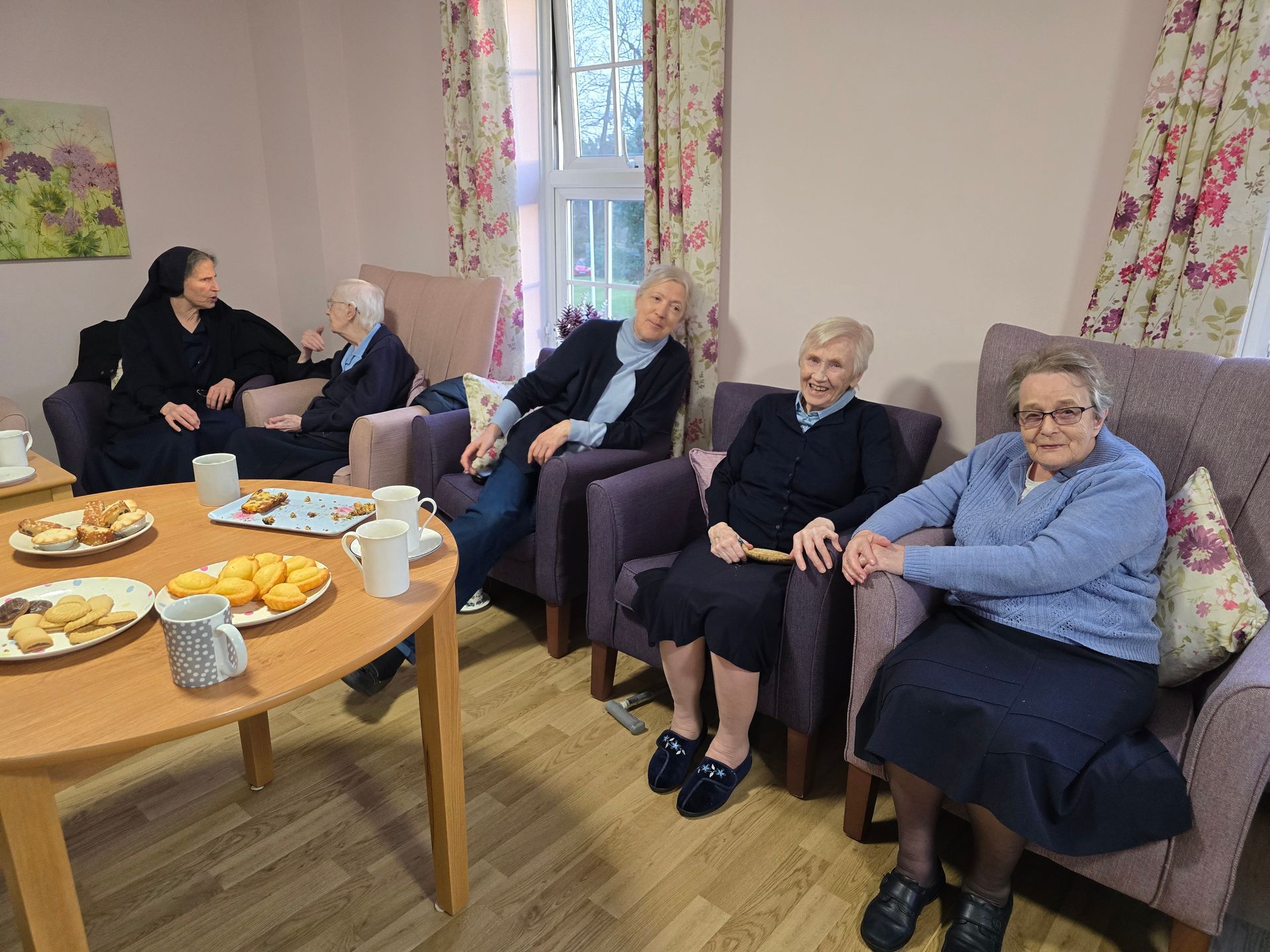 Group of people, some older, sitting in armchairs in a room. A table of snacks is in the foreground.