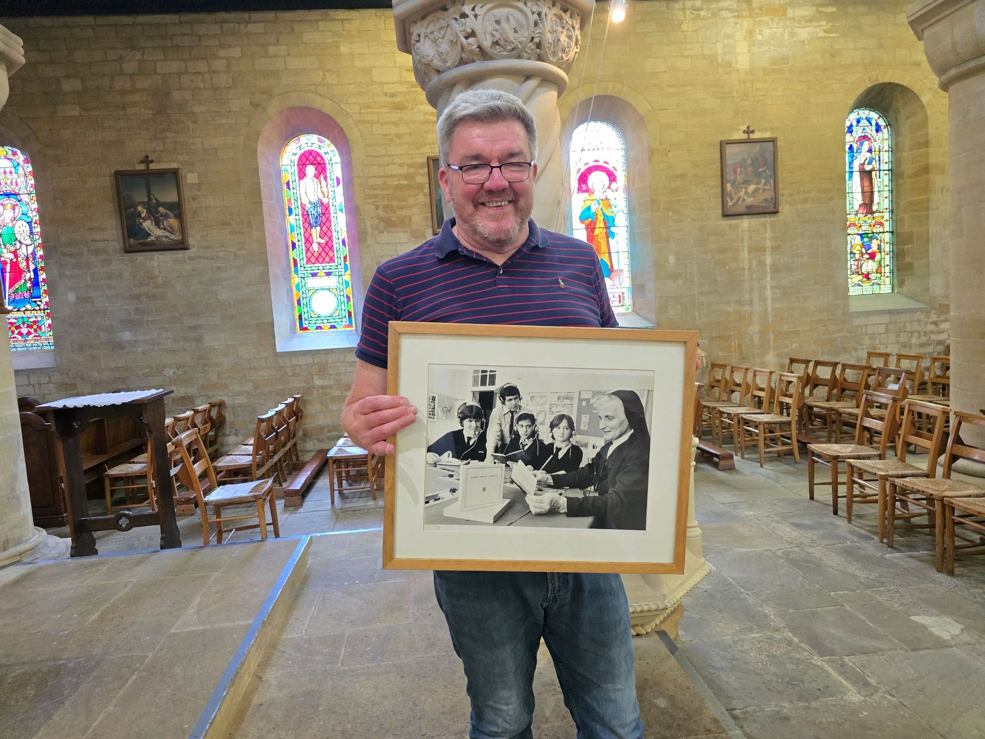 Man in church holds a framed black and white photo. He smiles. Colorful stained-glass windows in background.
