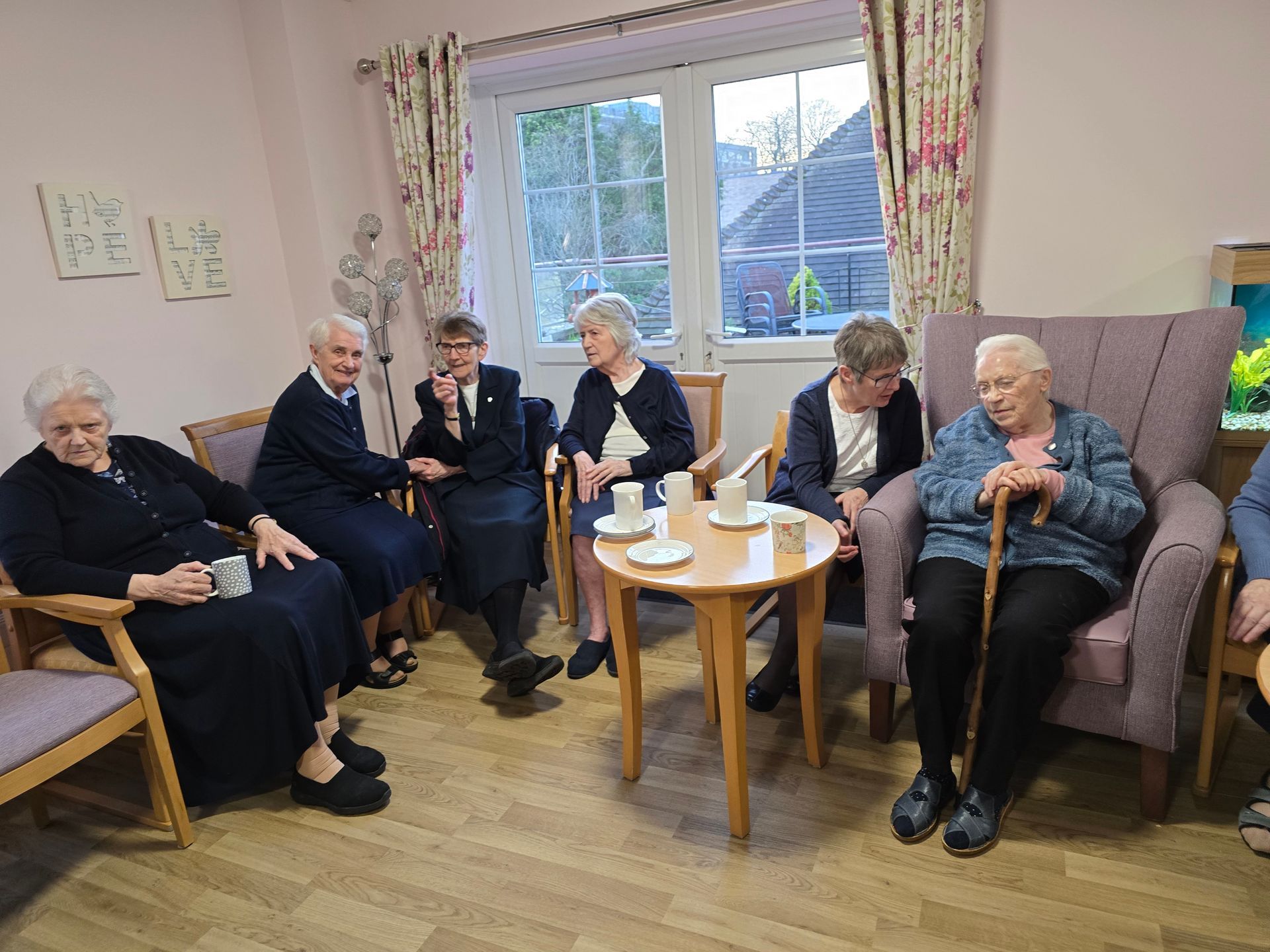 Group of older women seated, having tea indoors. Some hold mugs, one uses a cane, near a window.