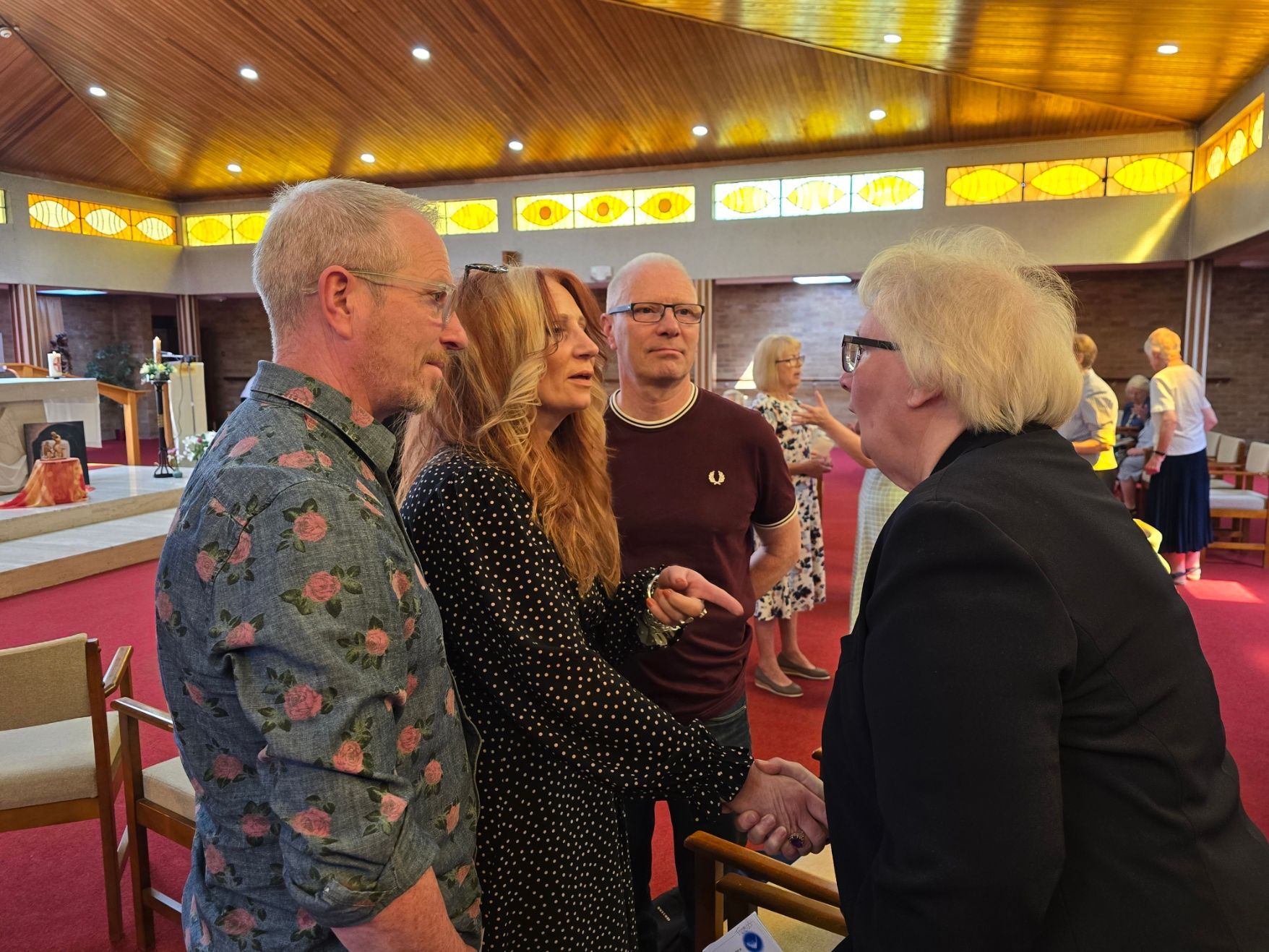 People in a church interior shake hands. Woman in black blazer shakes hands with woman in polka dots. Others nearby.
