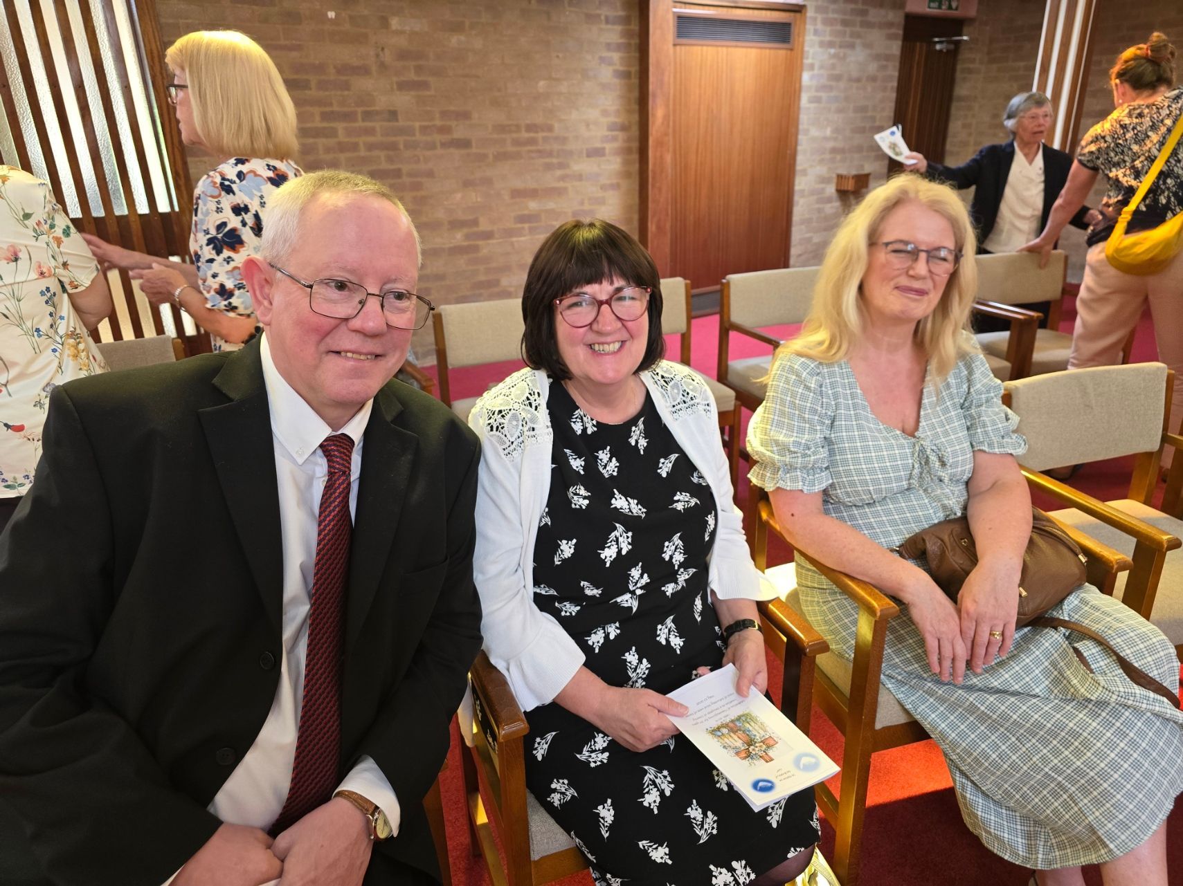Three people seated in a church. A man in a suit, a woman in a floral dress, and another woman, all smiling.