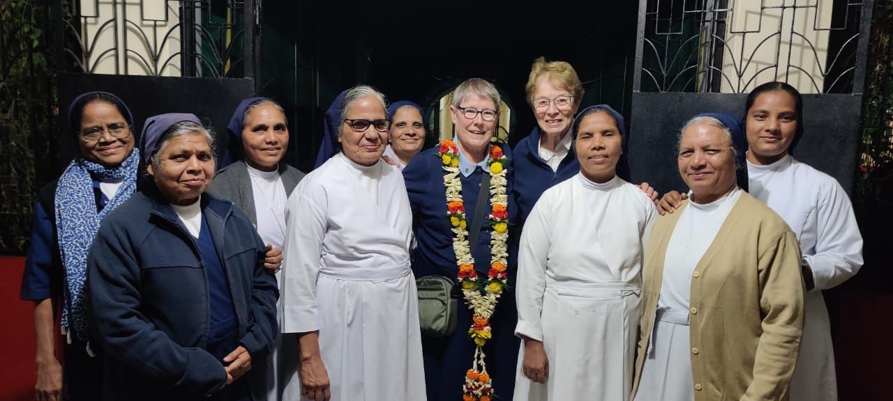 Group of nuns posing together outdoors, several wearing white habits, one has a garland.