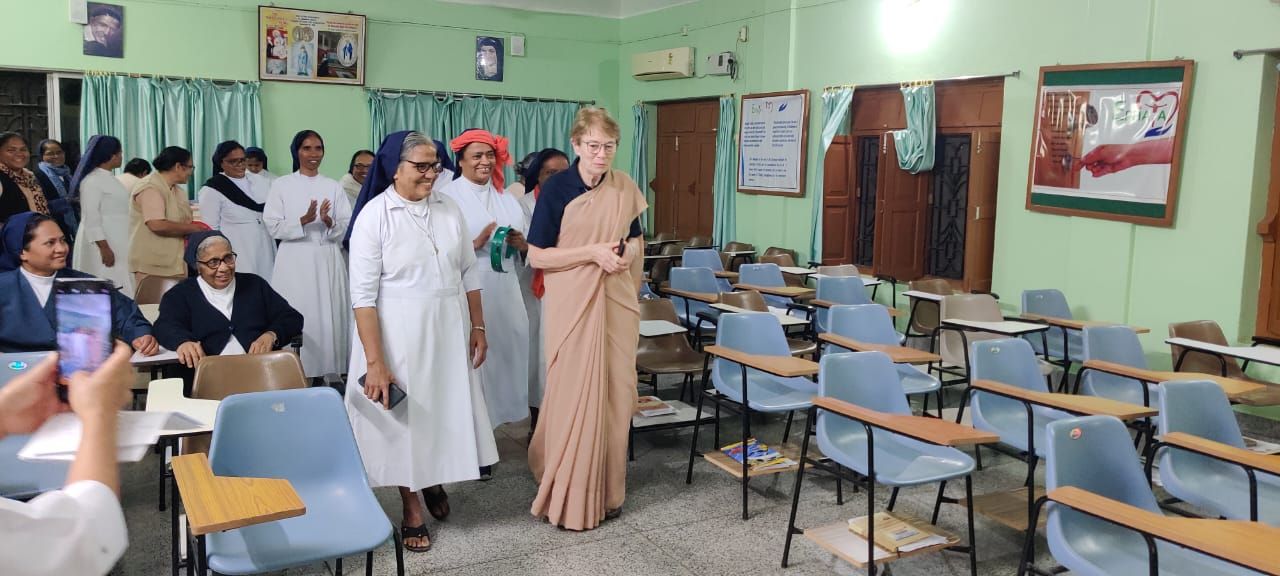 A woman in a saree walks through a classroom, followed by nuns. Others watch from the background.