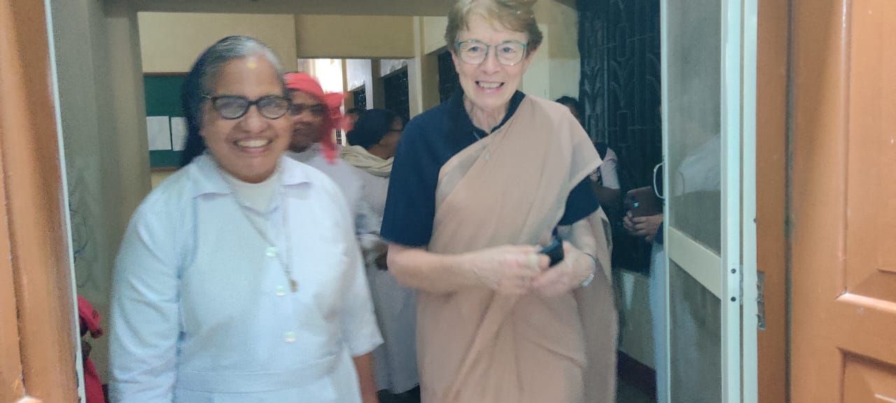 Two women smiling, one in a white habit, the other wearing glasses and a tan sari, in a doorway.