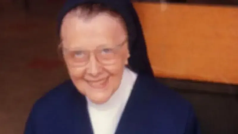 Nun in a blue habit smiles at the camera, wearing glasses, indoors, orange background.