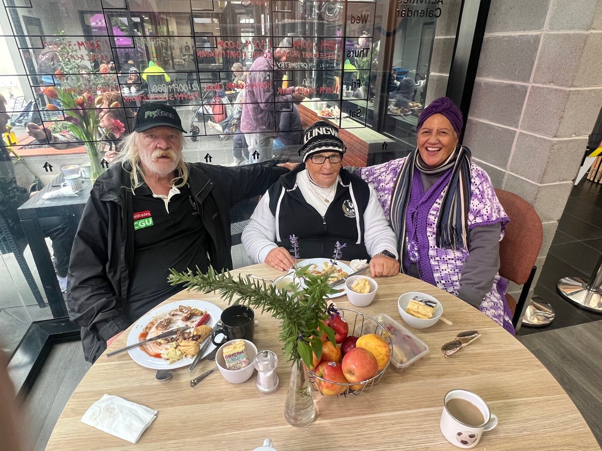 Three people smiling at a table in a cafe. A plate of food and fruit is visible.