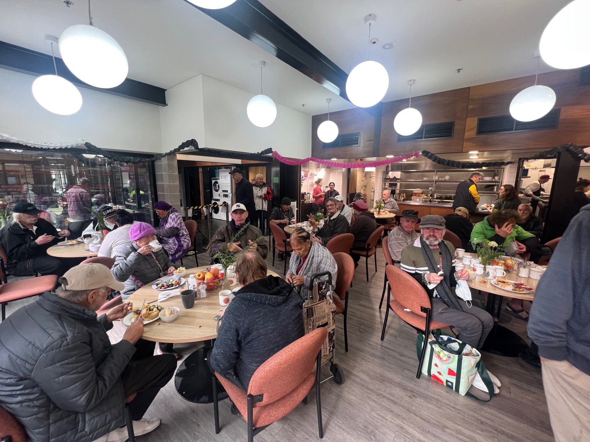People seated at tables in a dining area; some are eating, others talking. Round hanging lights above.