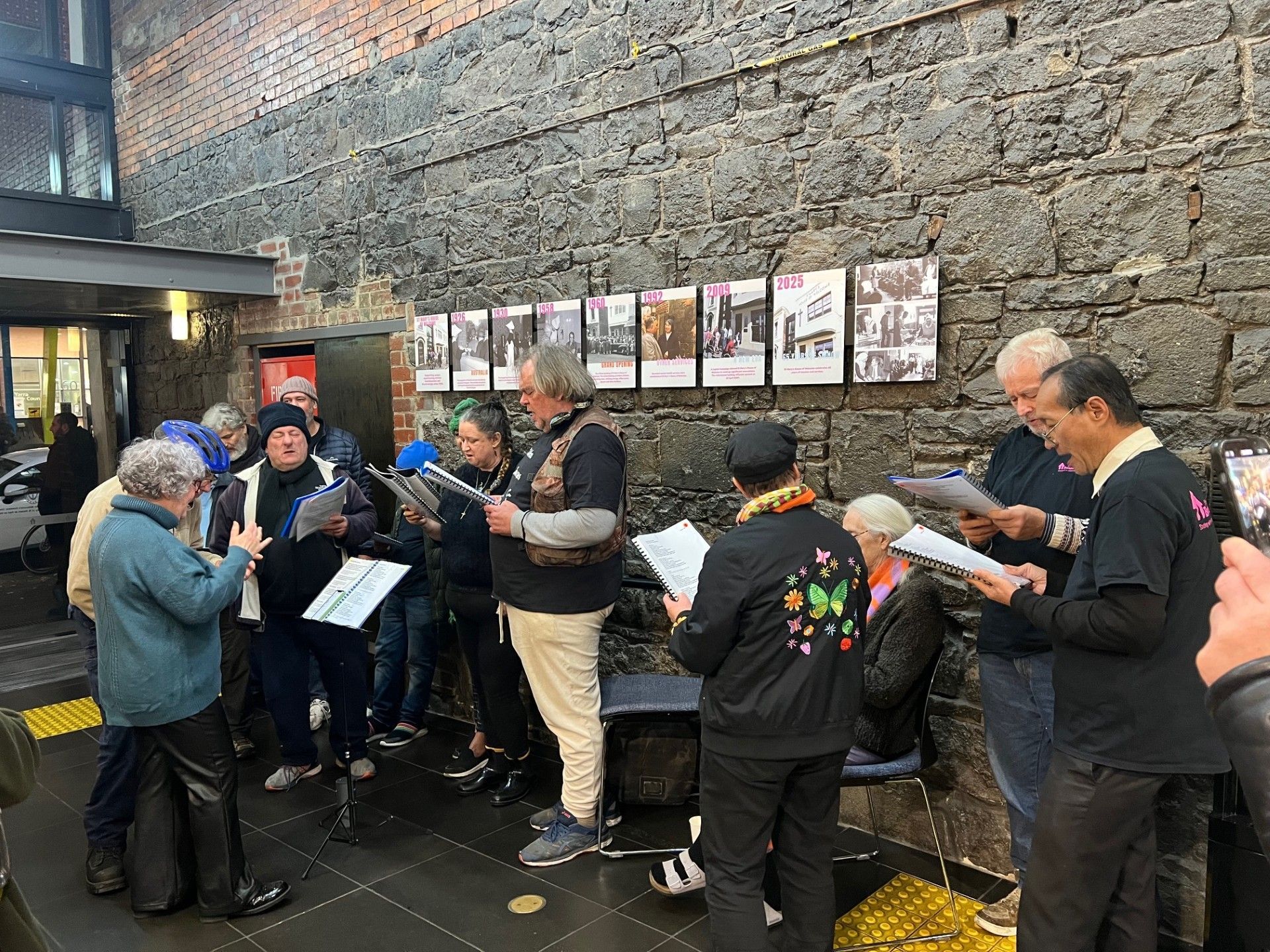 Group singing with sheet music, standing near a brick wall with photographs, indoors.
