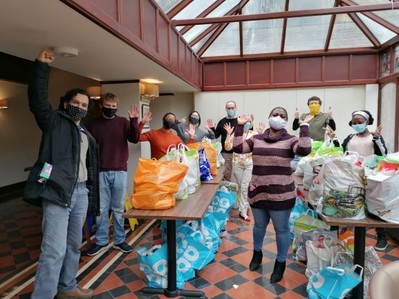 A group of people wearing masks are standing in a room with bags on tables.