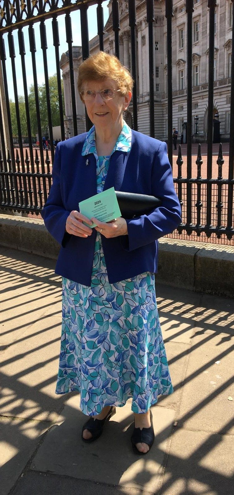 A woman in a blue jacket and blue dress is standing in front of a fence holding a piece of paper.