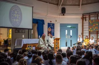A gathering with a bishop speaking at a podium to a crowd of schoolchildren; a screen displays a school crest.