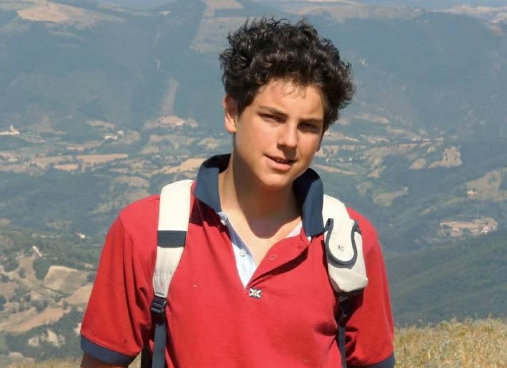 Carlo Acutis wearing a red shirt and a backpack is standing on top of a hill.