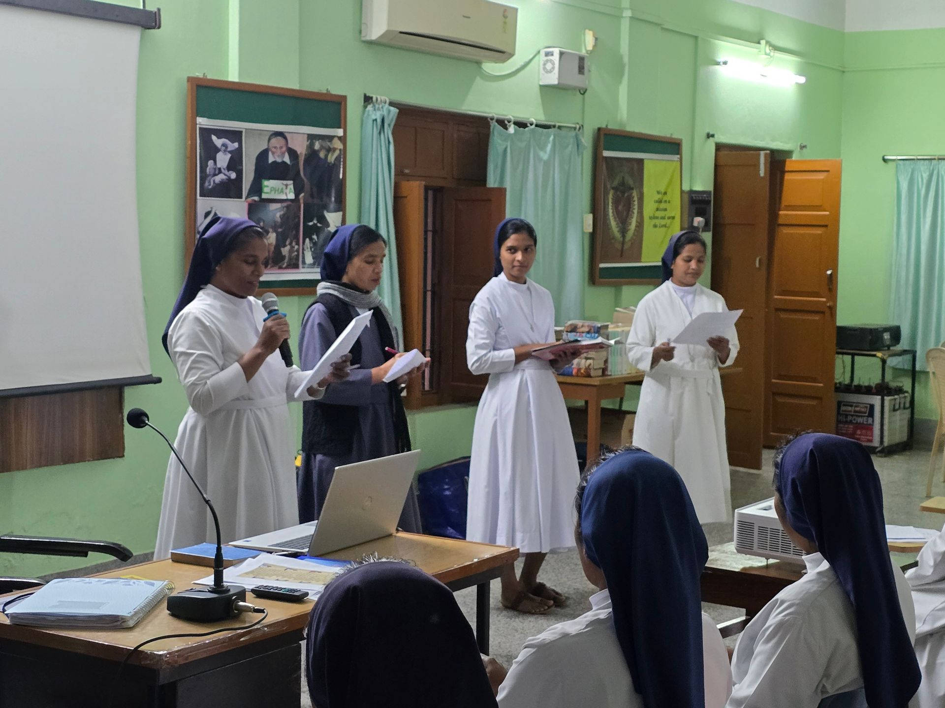 Nuns giving a presentation in a room. Four nuns stand at the front, speaking and holding papers. A laptop is visible.