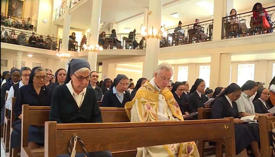 Priest in ornate vestments leads a service in a church. Nuns and worshippers attend.