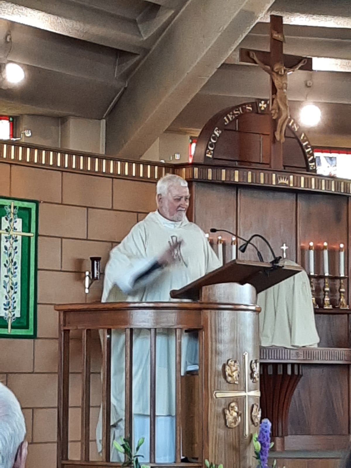 Priest in white robes at a wooden pulpit in a church, gesturing, with a crucifix and candles in the background.