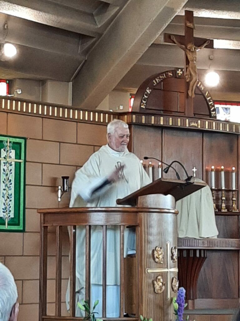 Clergyman in white robes at a wooden lectern, gesturing; crucifix behind, church interior.