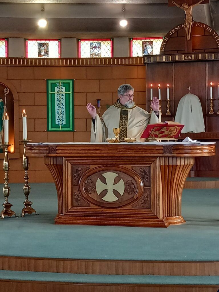 Priest at altar, raising hands during Mass. Brown wooden altar, stained glass windows, candles.