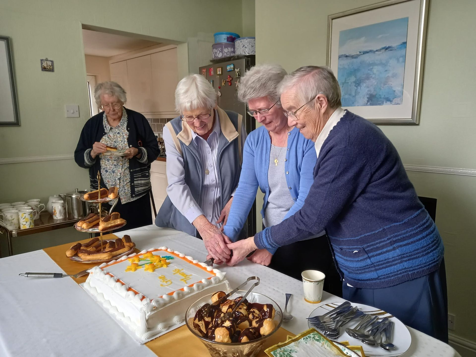 Four elderly women cutting a cake together at a table, celebrating. One woman is in the background, and the room has a homely setting.