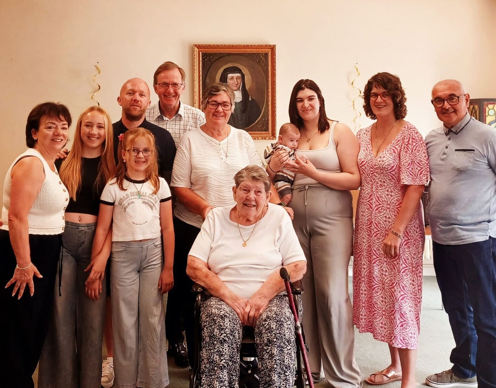 Group of people of all ages, mostly smiling, posing indoors with elderly woman seated in wheelchair.