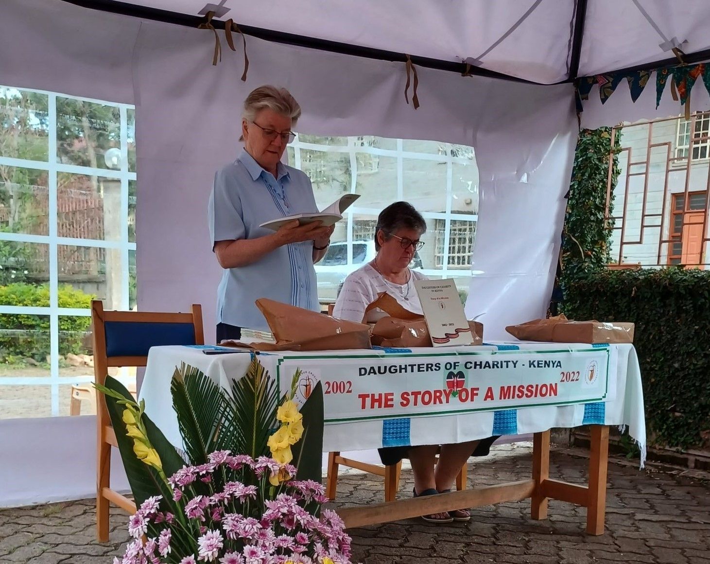 Two women read at a table under a tent. A banner reads,