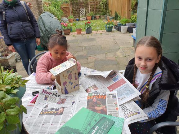 Two young girls are sitting at a table with newspapers and a box.