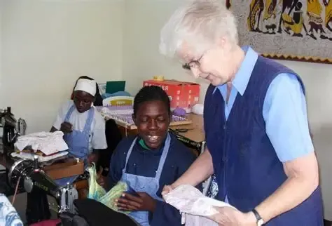 A woman in blue helps two others sew in a room with sewing machines.