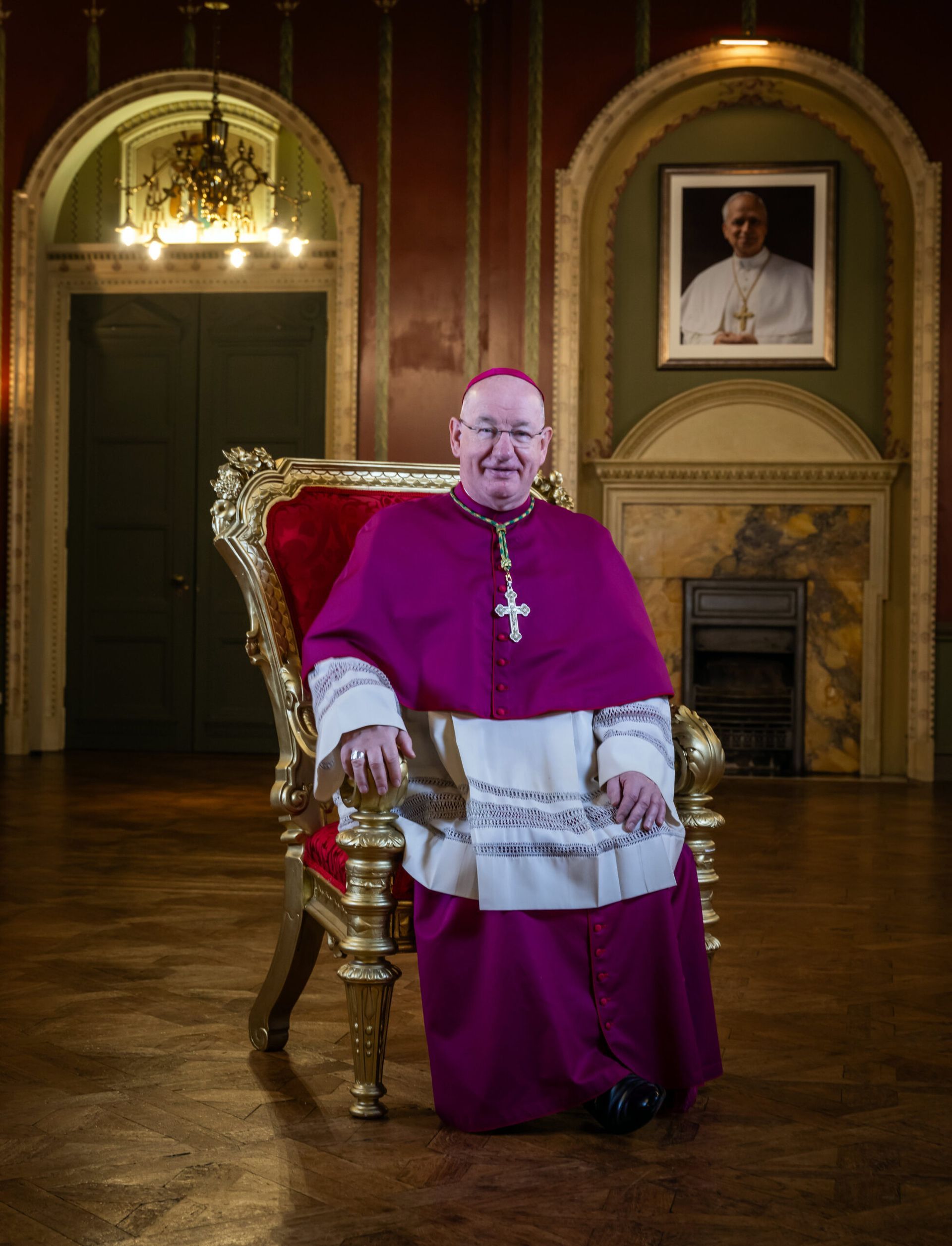 A bishop in purple and white vestments sits on an ornate gold chair in a room with a portrait of a pope on the wall.