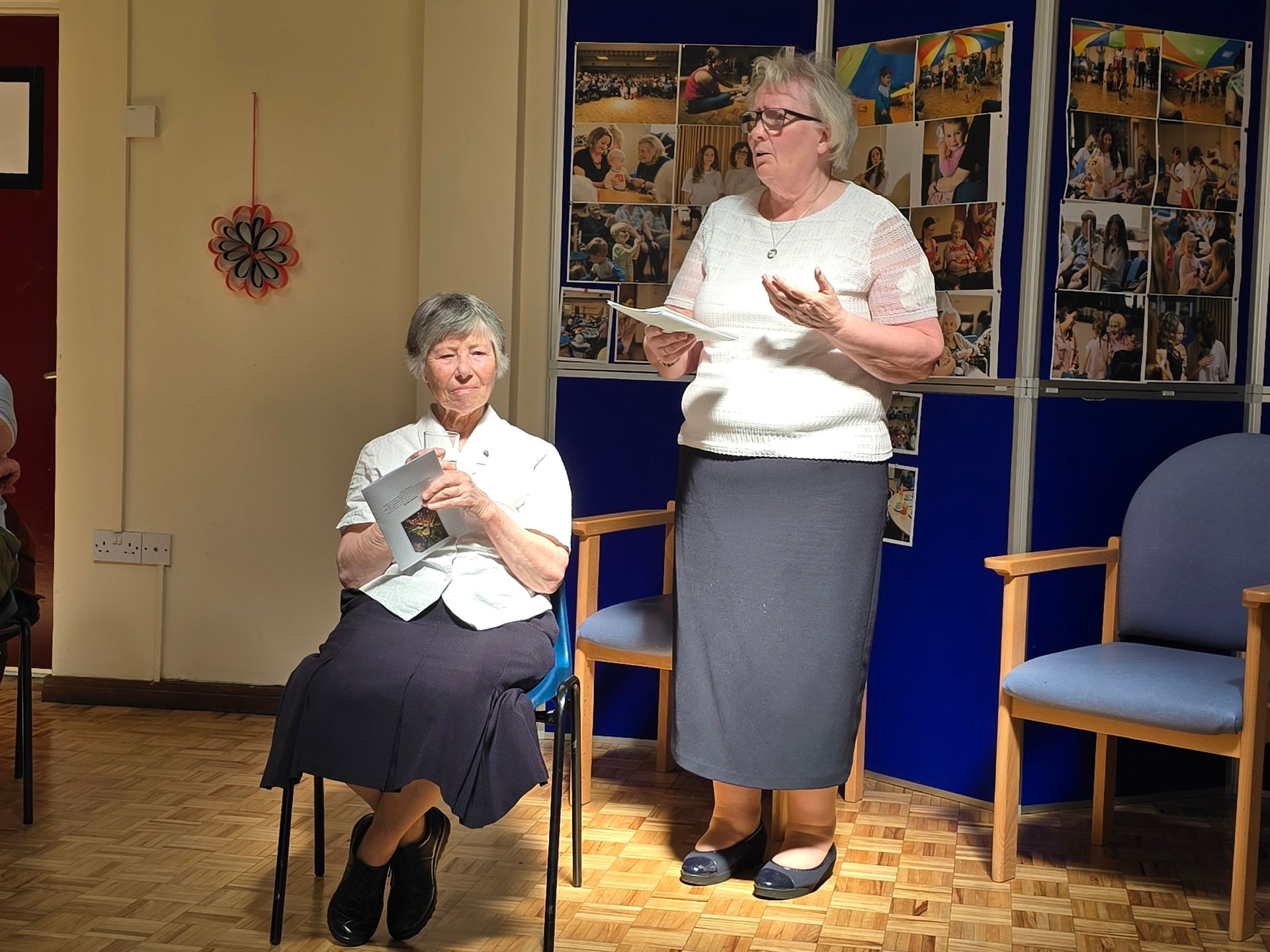 Two women at an event. One speaks, holding papers, the other sits with a book. Blue wall with photos.