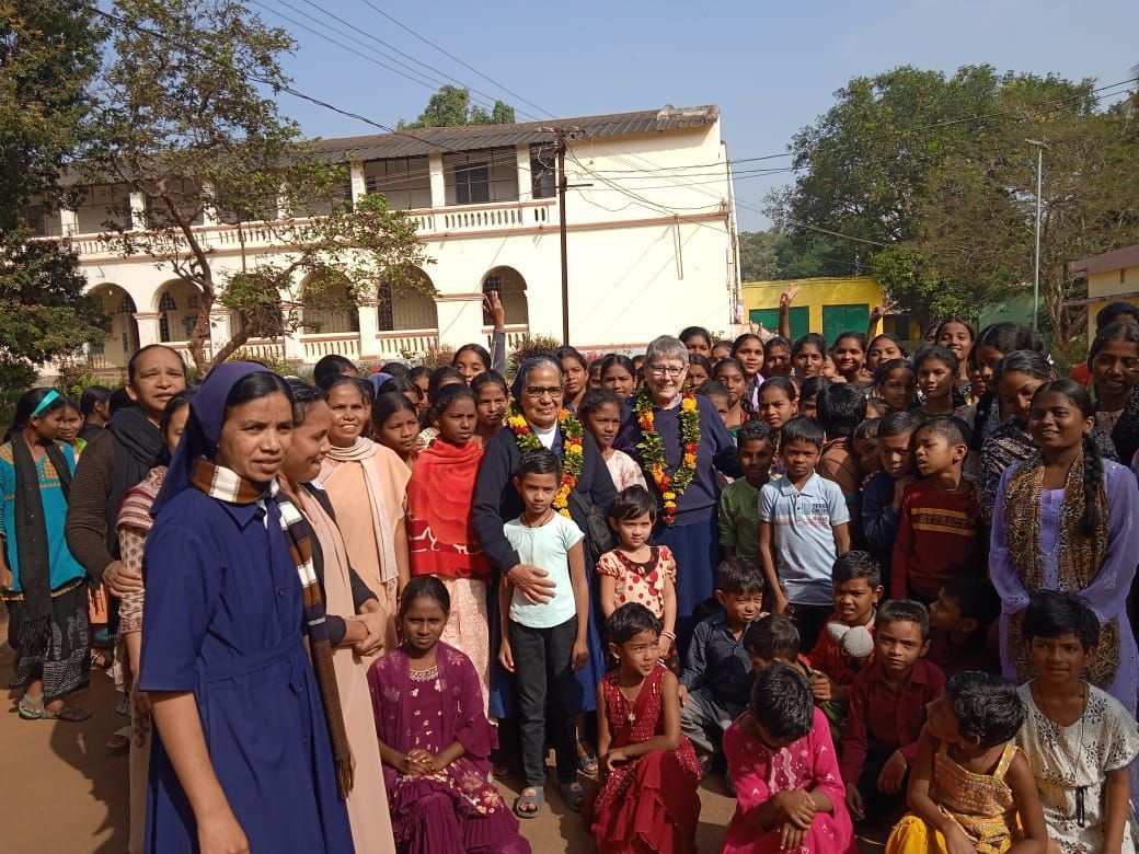 Group photo: children, nuns, and men in front of a building. Sunny day. People wearing flower garlands.